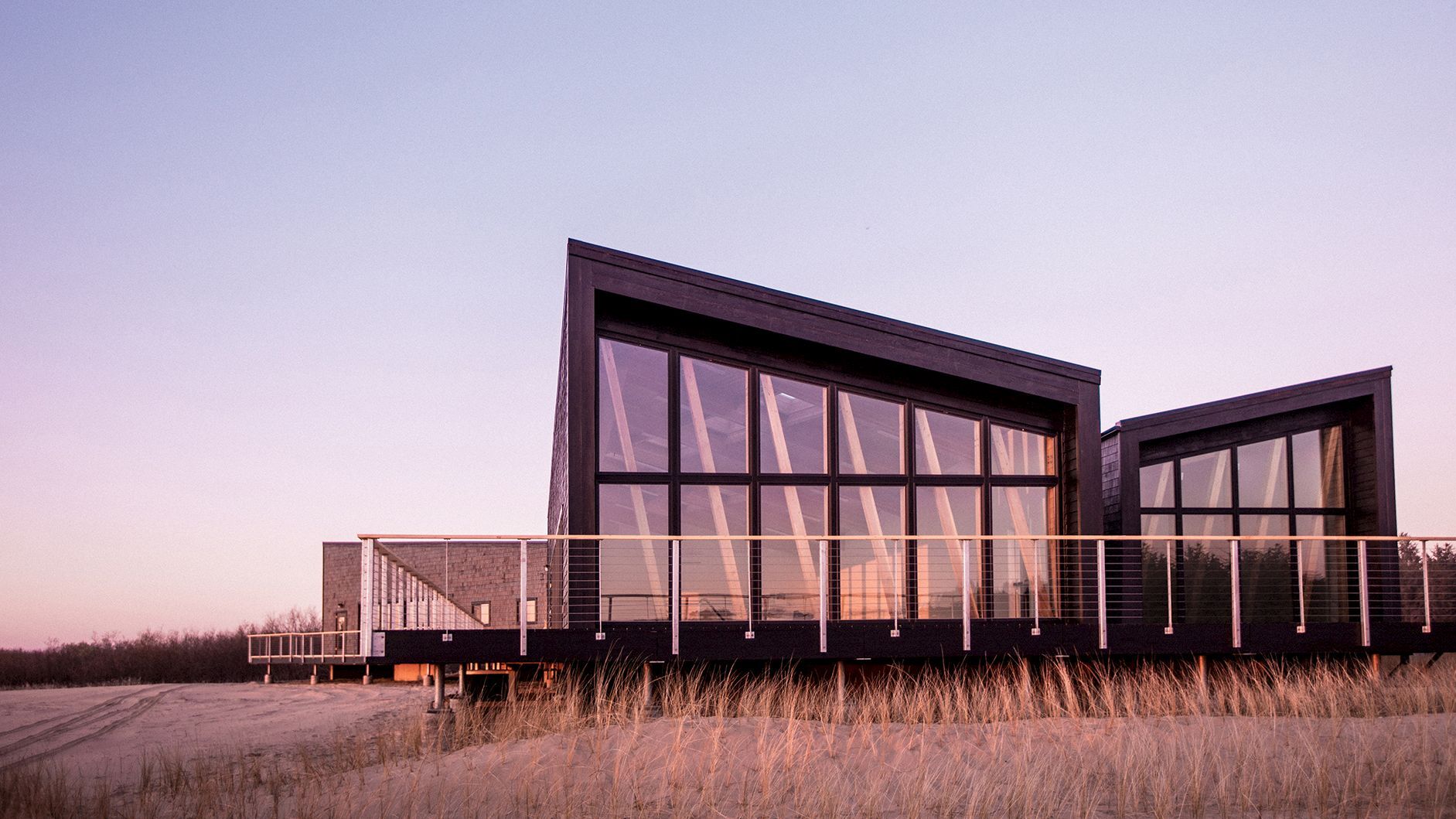The image shows a modern building with large windows and a slanted roof, set against a sunset sky, surrounded by tall grass.