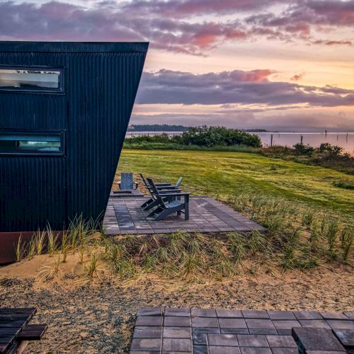 A modern black building with large windows overlooks a grassy field and ocean at sunset, featuring a wooden patio with chairs.