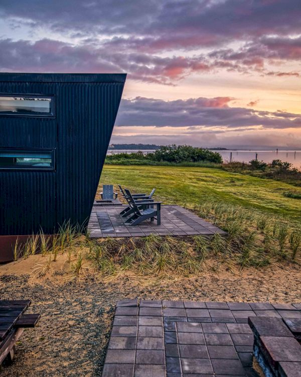 A modern black building with large windows overlooks a grassy field and ocean at sunset, featuring a wooden patio with chairs.