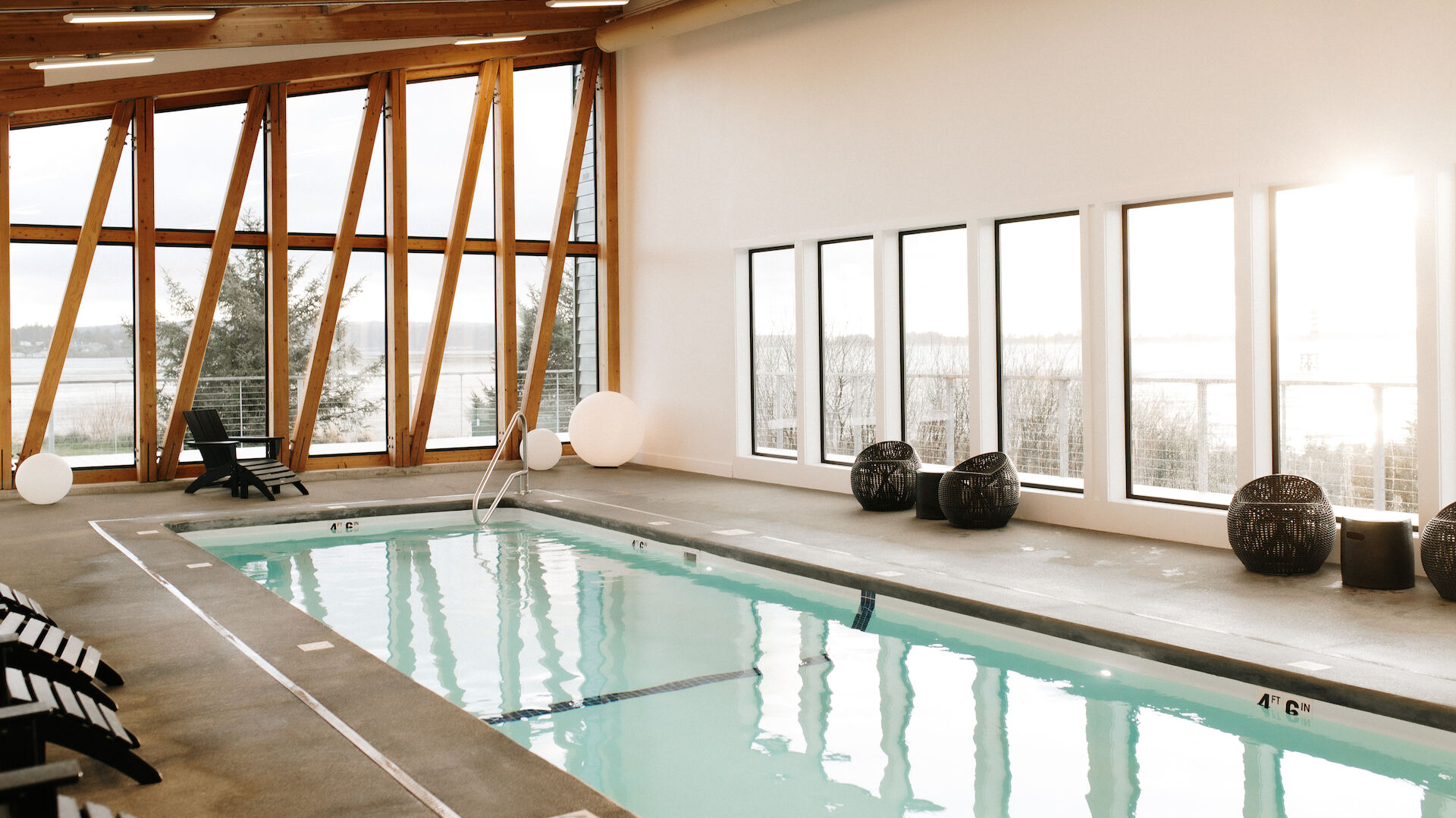 A modern indoor pool with wooden beams, lounge chairs, large windows, and spherical lights, offering a serene and stylish atmosphere.