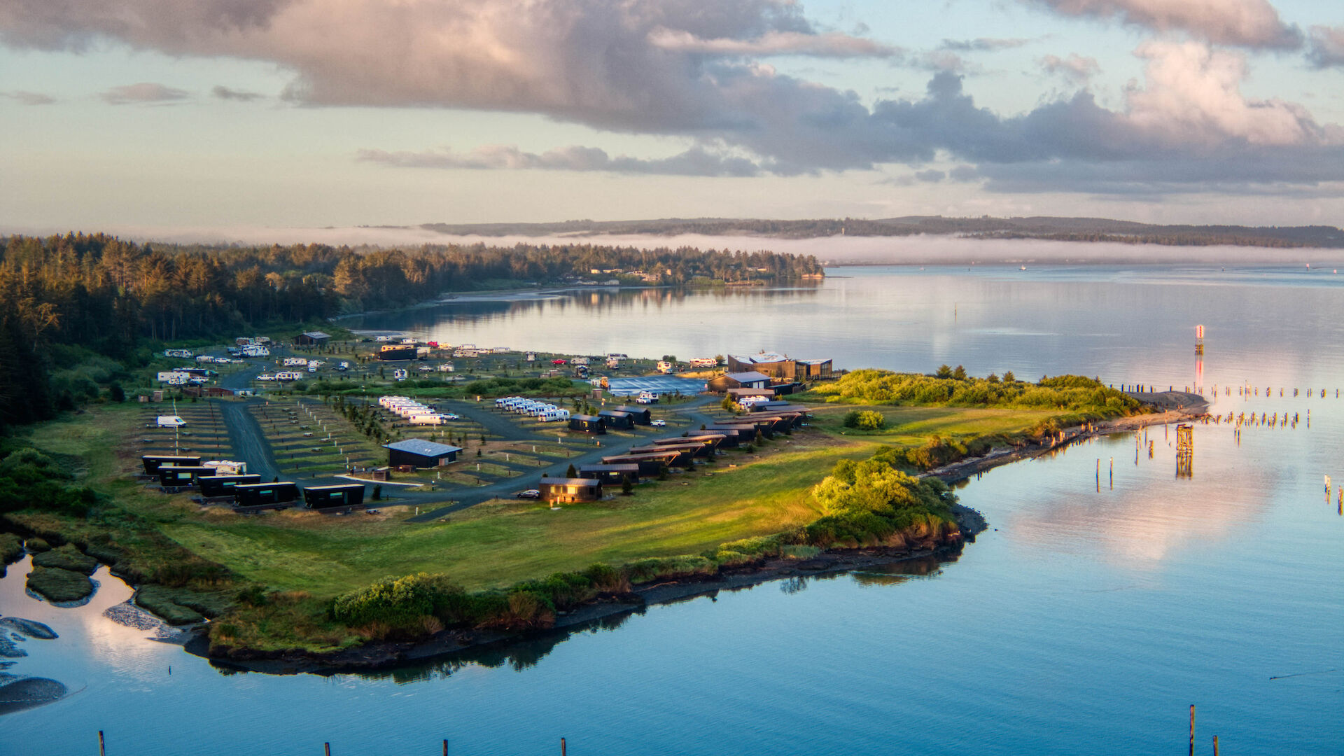 A coastal landscape with a grassy peninsula, buildings, RVs, and calm water reflecting the sky and clouds at sunset.