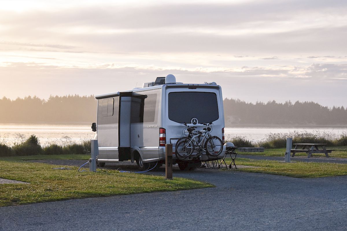A camper van parked by a lakeside at sunset with bicycles attached to the rear. A picnic table and grassy area are visible nearby.