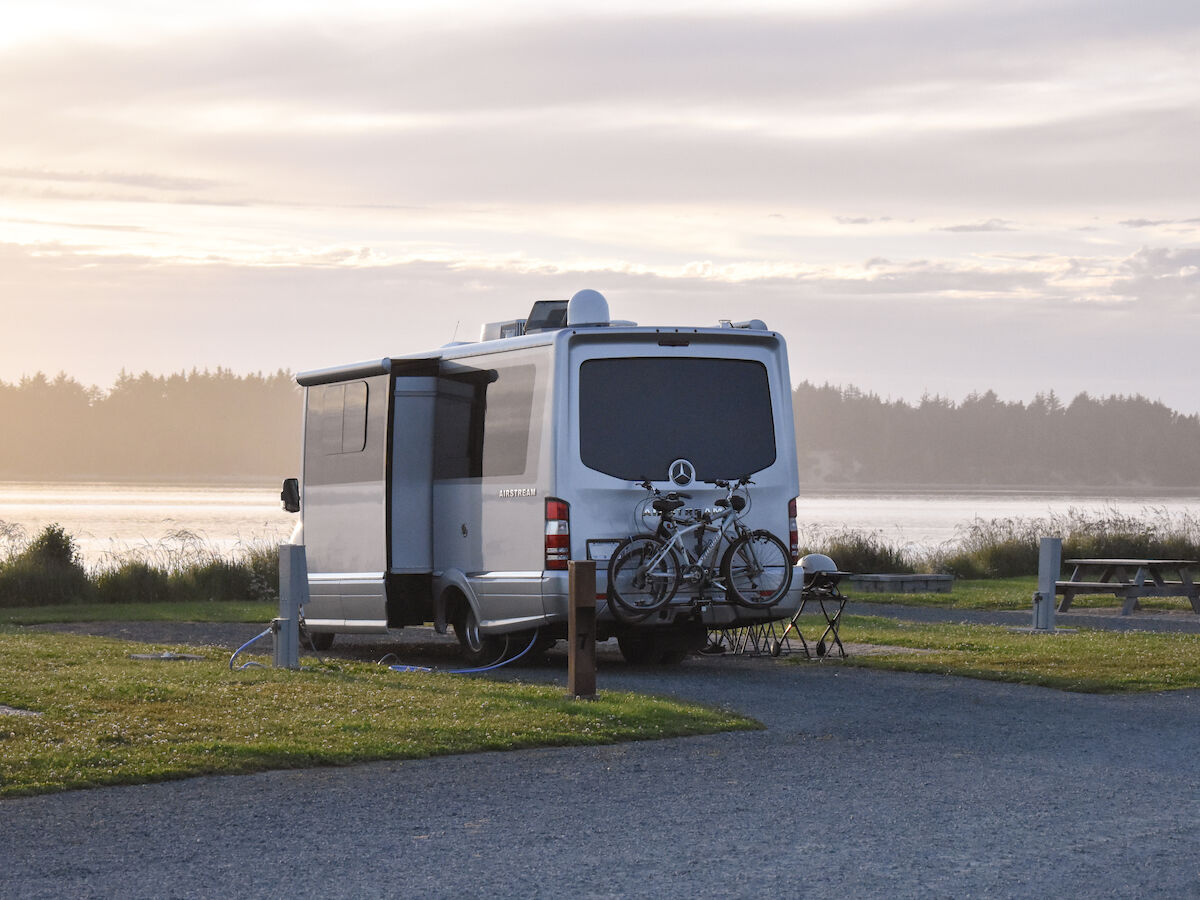 A camper van parked by a lakeside at sunset with bicycles attached to the rear. A picnic table and grassy area are visible nearby.