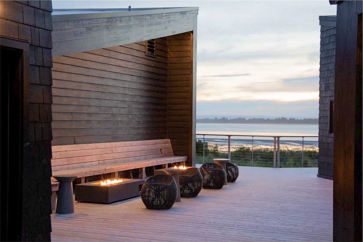 A modern outdoor seating area with wooden benches, decorative lanterns, and a view of the water in the background at dusk.