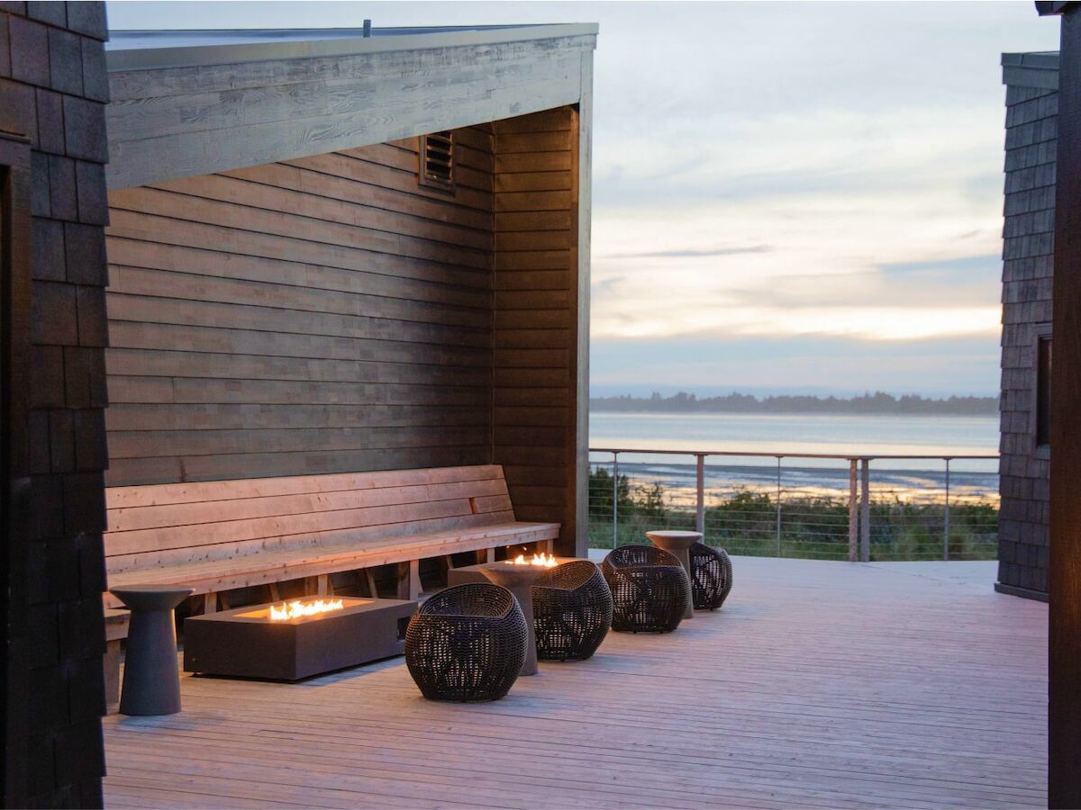 A modern outdoor seating area with wooden benches, decorative lanterns, and a view of the water in the background at dusk.