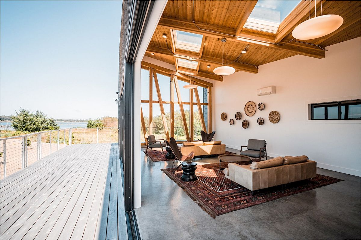 A modern living room with wooden ceiling, skylights, and outdoor view. Features cozy sofas, circular wall decor, and a spacious deck outside.
