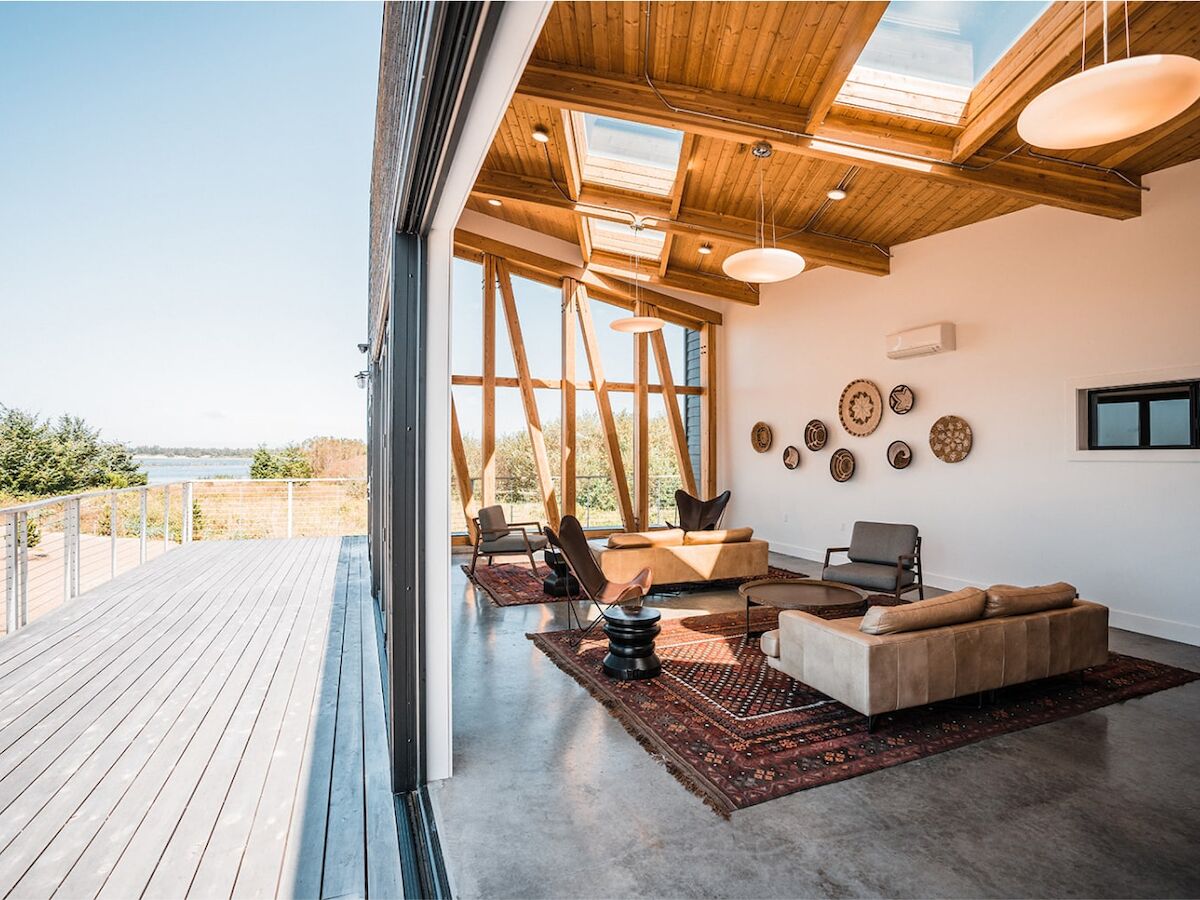 A modern living room with wooden ceiling, skylights, and outdoor view. Features cozy sofas, circular wall decor, and a spacious deck outside.