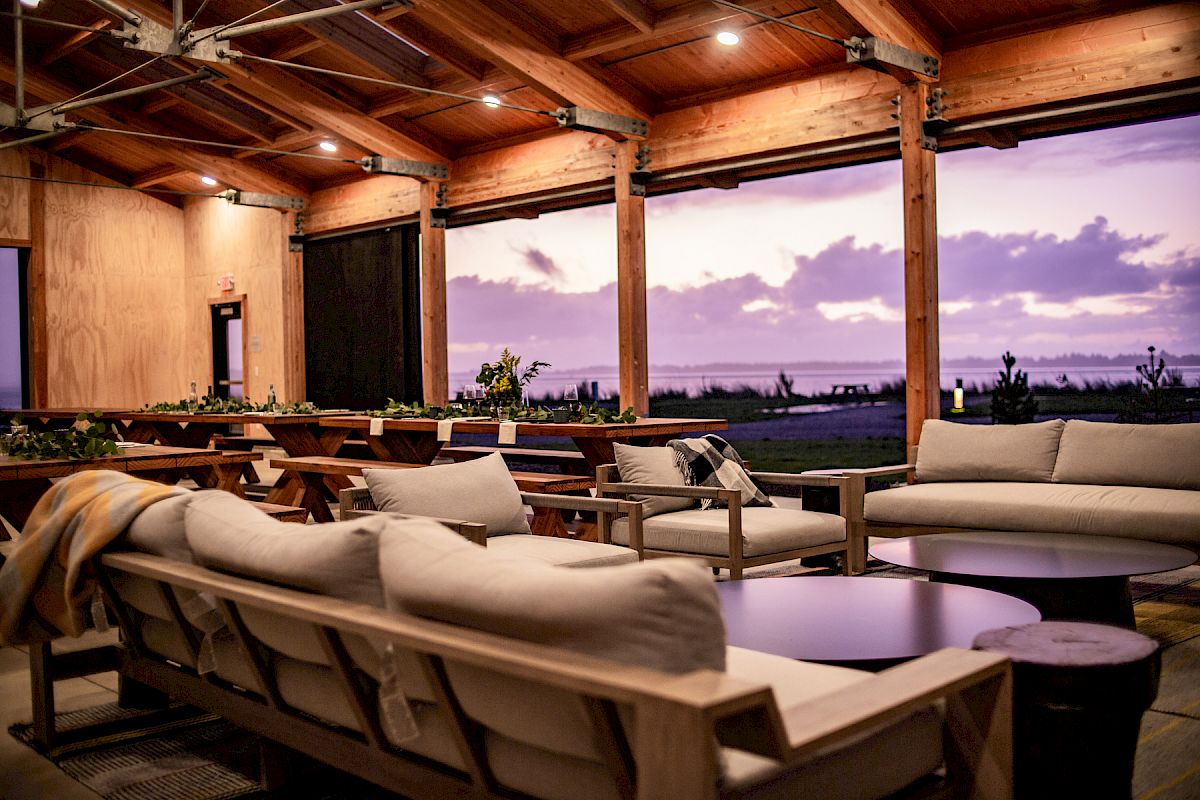 A cozy outdoor lounge with comfortable sofas and a wooden ceiling, overlooking a scenic sunset view through large open windows.
