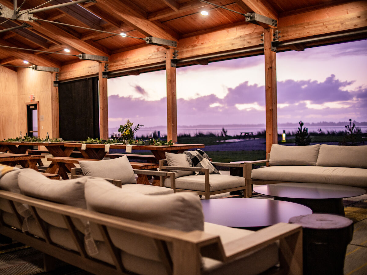 A cozy outdoor lounge with comfortable sofas and a wooden ceiling, overlooking a scenic sunset view through large open windows.