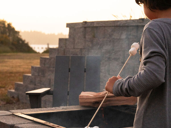 A child wearing a gray sweater stands by a concrete grill, roasting marshmallows on a stick at sunset.