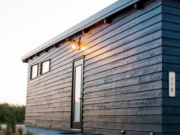A small, modern, black wooden house with a slanted roof and a lit exterior light, located on a sandy landscape with a gravel path.