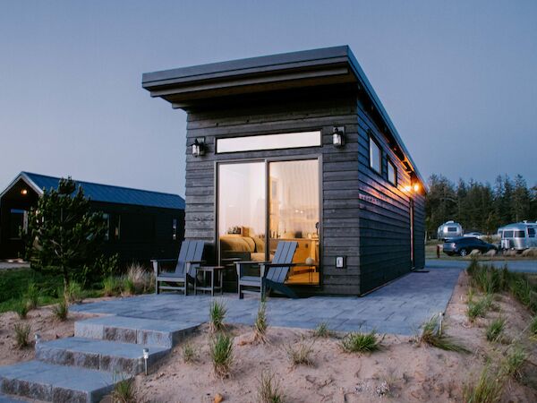 A modern tiny house with large windows, patio chairs, and steps leading up to it; nearby trees and trailers are visible in the background.