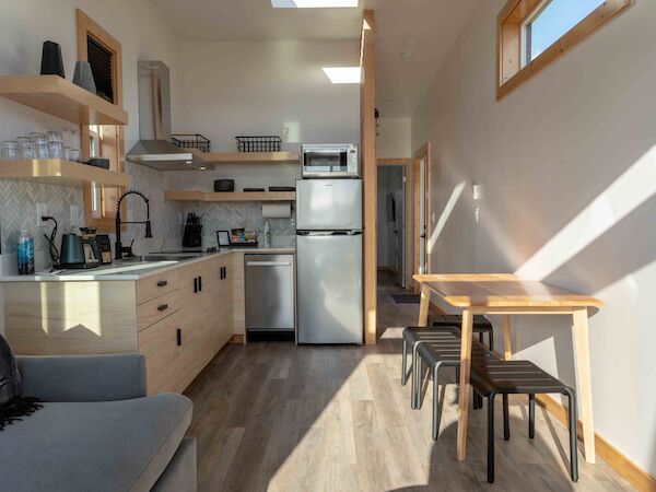 A modern kitchen and dining area with wooden cabinets, stainless steel appliances, a table with stools, and a sofa. Sunlight streams in.