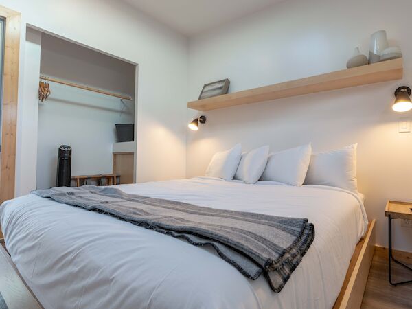 A cozy bedroom with a double bed, white linens, a striped throw, a shelf with decor, and a small side table is shown.
