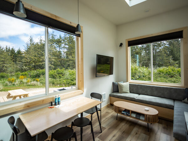 The image shows a cozy room with large windows, a dining table, chairs, a wall-mounted TV, and a comfortable seating area with outdoor views.