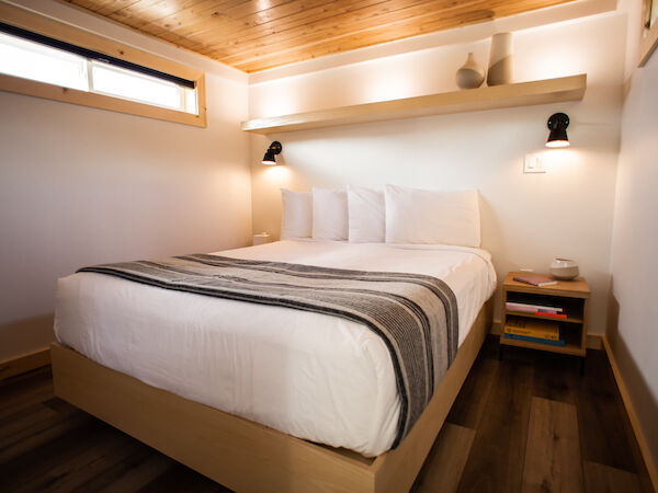 A cozy bedroom features a neatly made bed with a striped blanket, wooden accents, a small shelf, and warm lighting from wall sconces.