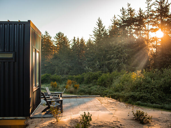 A modern black cabin with patio chairs is surrounded by lush greenery and trees, basking in a beautiful sunrise.