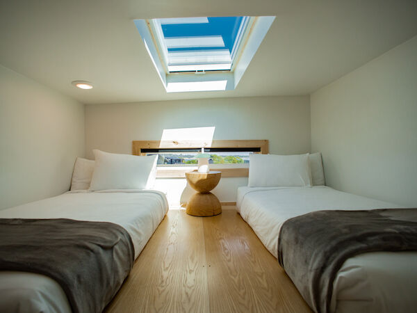 A bright, compact bedroom with two single beds, white linens, a small central stool, and a skylight letting in natural light. End.