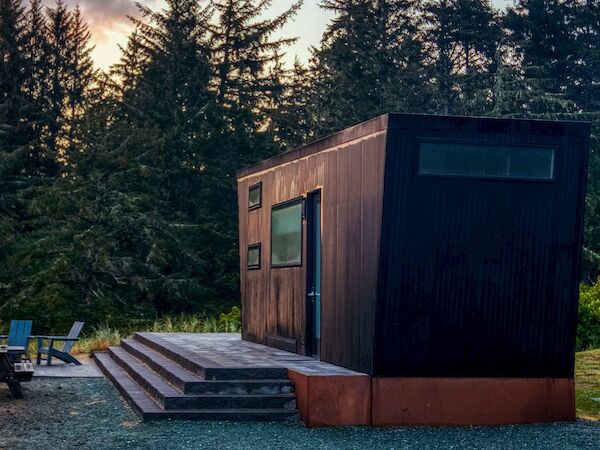 A modern cabin with steps is in a forest setting. Nearby, there's a picnic table and chairs on a deck at sunset.
