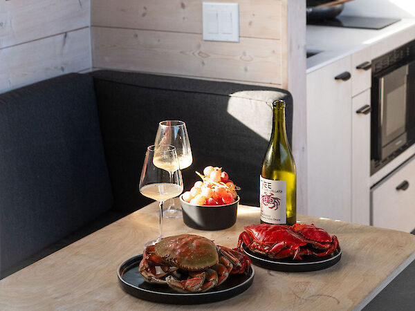 Cozy camper kitchen with a small table set for two: burger, steak, wine, and a bowl of snacks, chrome faucet, window light, compact stove, fridge.