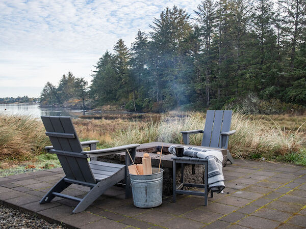 A lakeside outdoor seating setup with two Adirondack chairs around a small fire pit on a stone patio, a bucket of firewood, and a forested shoreline in the background.