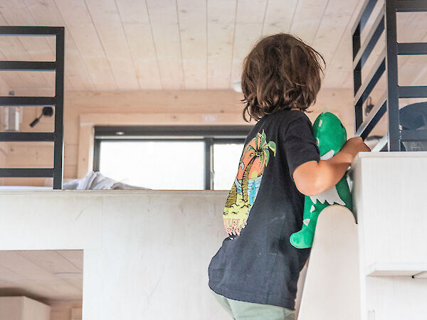 A child climbs a white wall with a green skateboard, wearing a black shirt and green shorts, inside a bright, airy room with wooden ceiling.