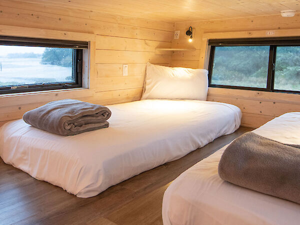 A cozy wood-clad bedroom with two twin beds, each with white bedding and a folded blanket, warm lighting, and large windows.