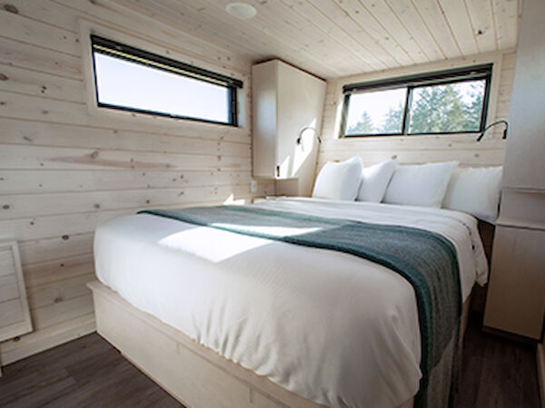 A cozy bedroom with wood paneling features a neatly made bed, two windows, and built-in lighting, creating a warm and inviting space.