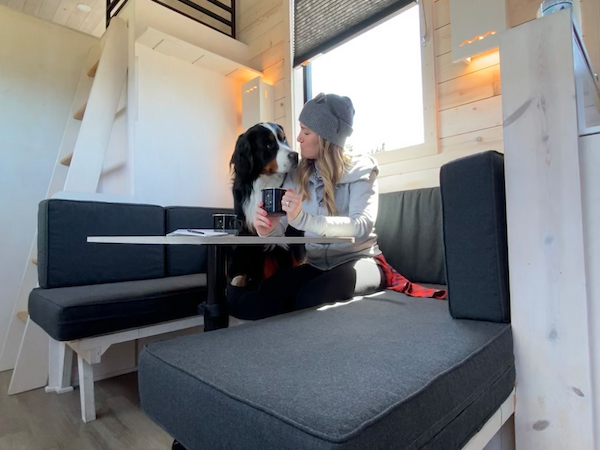 A person and a dog are sitting at a small table in a cozy, modern space with lofted beds and wooden walls. The scene is well-lit.