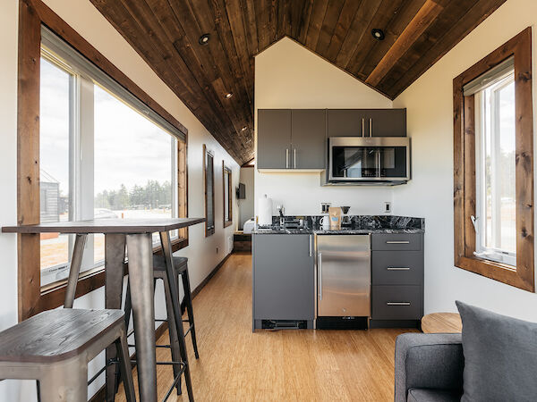 Modern tiny house interior with a small kitchen, wood accents, and windows. High table with stools and a cozy sitting area are visible.