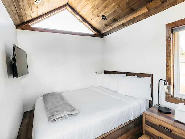 A cozy bedroom features a bed, a mounted TV, and a window, all under a wooden ceiling with a triangular skylight.