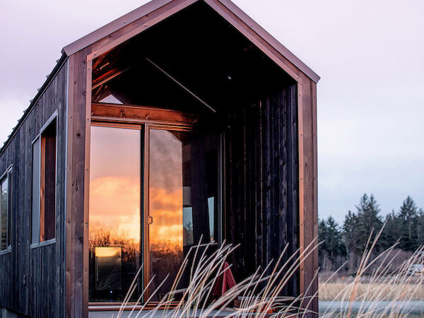 A small, modern cabin with a wooden exterior shows a sunset reflected in its glass door, surrounded by tall grasses and distant trees.