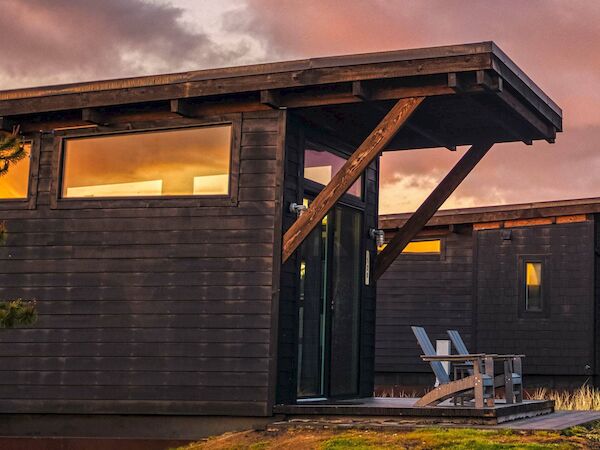 A modern wooden cabin with large windows, featuring two chairs on a small porch, set against a sunset sky.