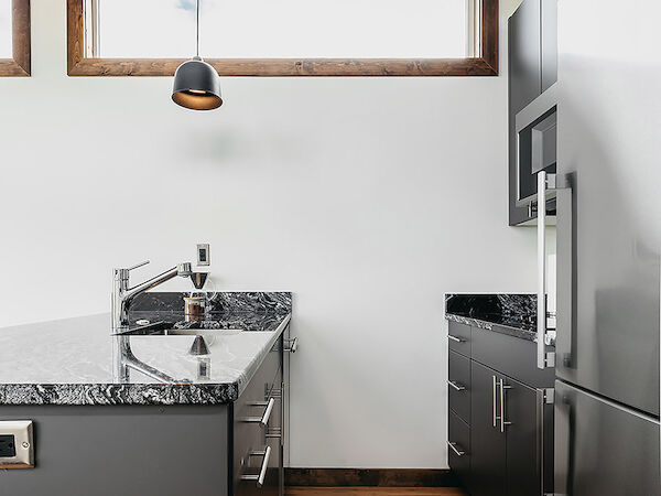 A modern kitchen with grey cabinets, marble countertops, stainless steel appliances, wooden flooring, and pendant lighting hangs from wooden beams.