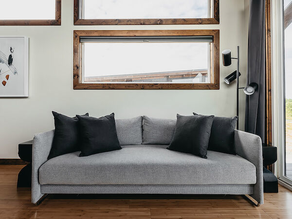 The image shows a modern living room with a gray sofa, black cushions, framed artwork, large windows, and a floor lamp against a wooden floor.