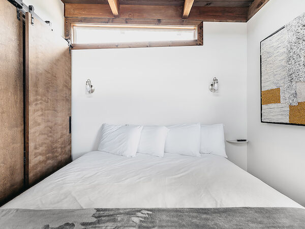 A cozy bedroom with a wooden ceiling, white walls, a bed with white linens, wall lights, and abstract artwork on the wall.