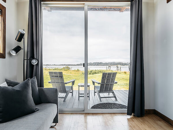 A cozy room with a sofa leads to a patio through sliding glass doors, featuring two chairs and a scenic view outside.