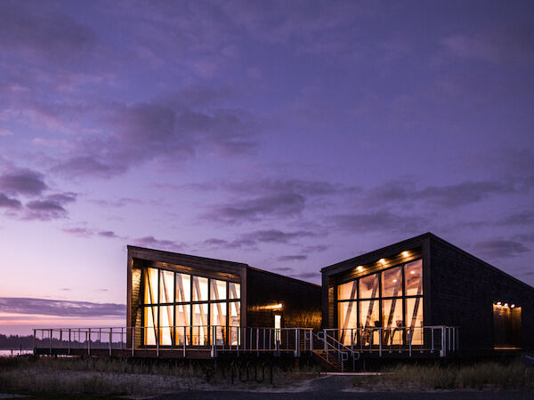 A modern building with large windows is illuminated under a purple sky at dusk, featuring a scenic outdoor view and warm interior lights.