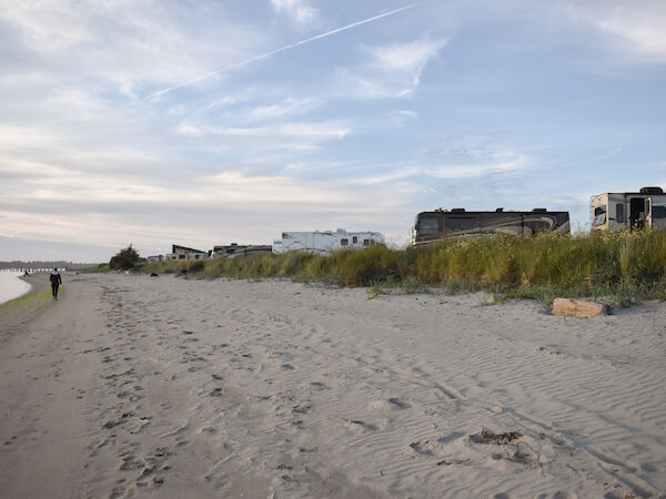 A sandy beach with footprints leads to several RVs parked near dunes, with grass and a cloudy sky in the background.