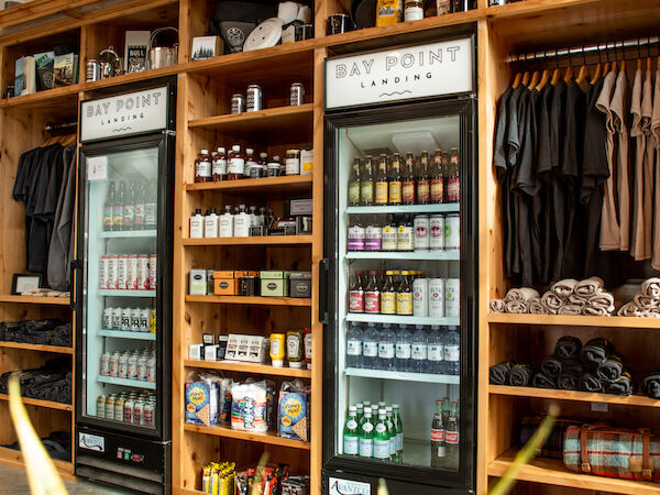 The image shows a shop interior with wooden shelves, refrigerators stocked with drinks, merchandise, and a plant in the foreground.