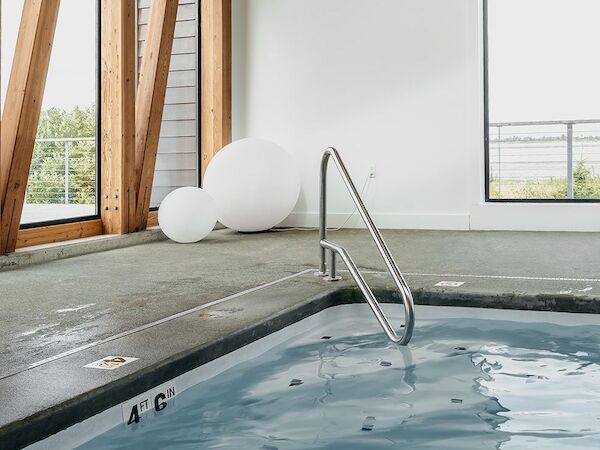 The image shows an indoor pool area with a handrail, windows, and modern wooden beams. Large white spherical decor is seen in the corner.
