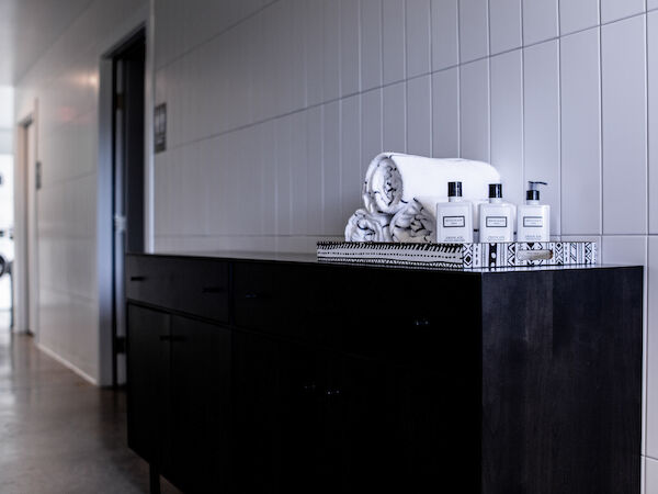 A hallway with a dark cabinet topped with rolled towels and toiletries. The background shows a partially visible laundry room with machines.