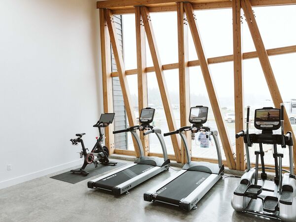The image shows a gym with a stationary bike, two treadmills, and an elliptical machine near large windows.