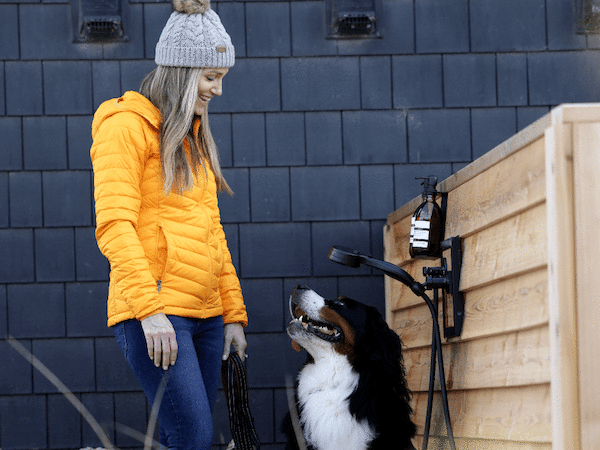 A person in an orange jacket and beanie stands beside a large dog outside near a wooden wall and outdoor shower.