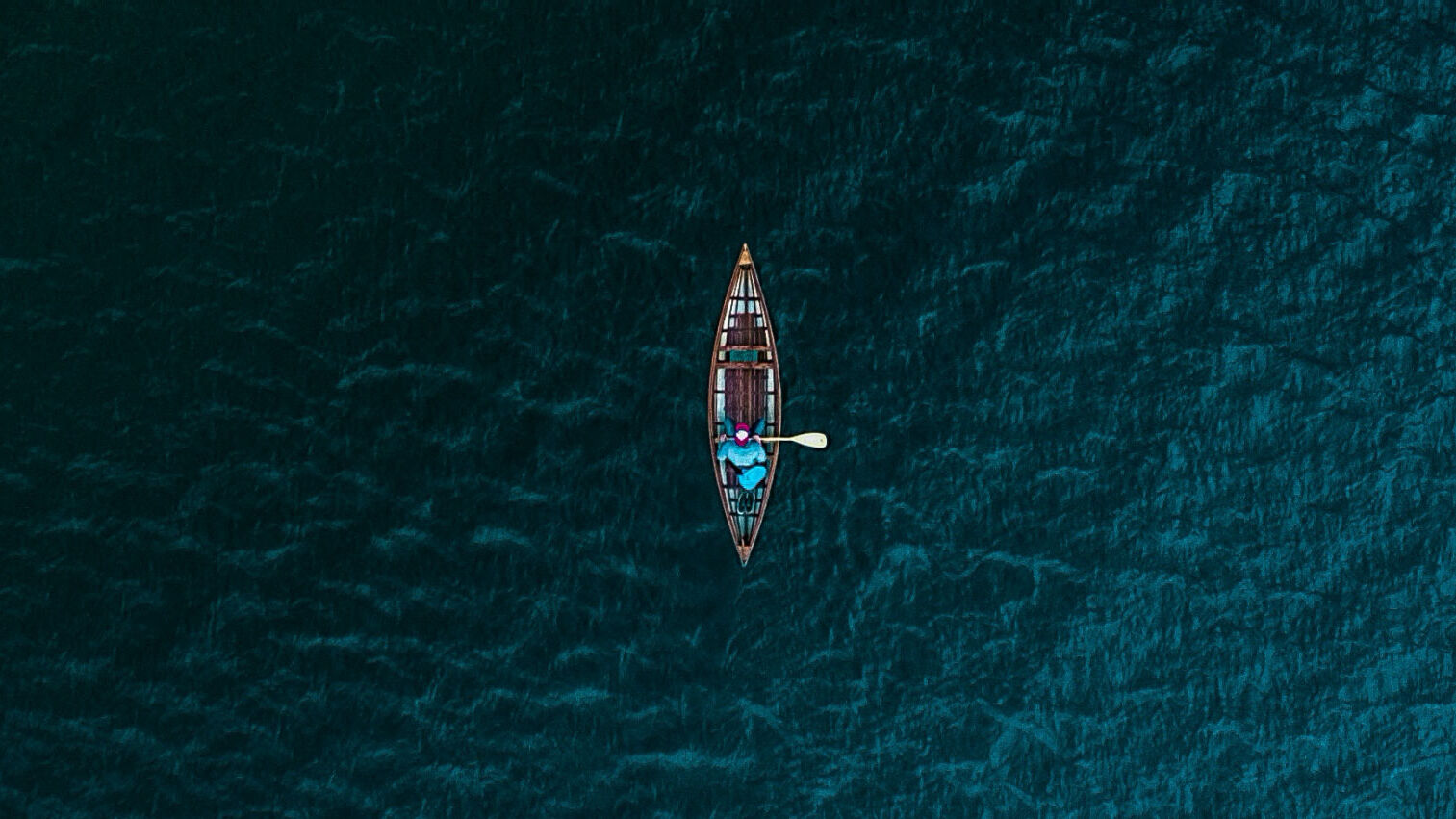 Aerial view of a lone canoeist in a small boat on a vast, dark blue body of water.
