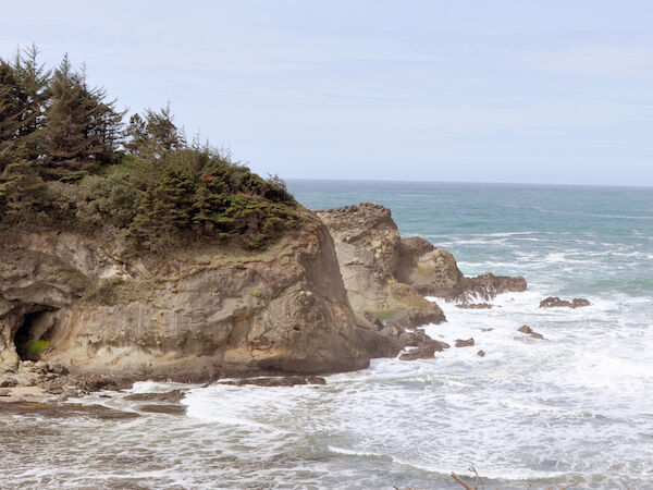 A coastal scene with rocky cliffs, waves hitting the shore, and lush greenery in the foreground under a clear sky.