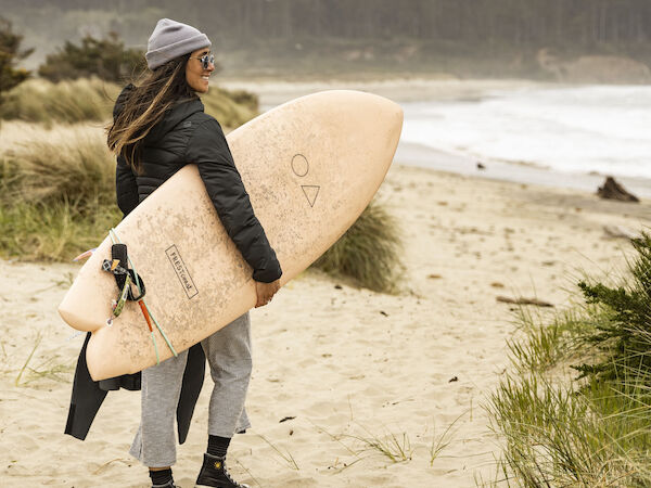 A person stands on a sandy beach holding a surfboard, wearing casual outdoor attire. The ocean and forested area are in the background.