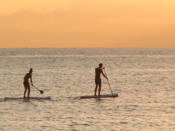 Two people are paddleboarding on the ocean at sunset with an orange sky in the background.