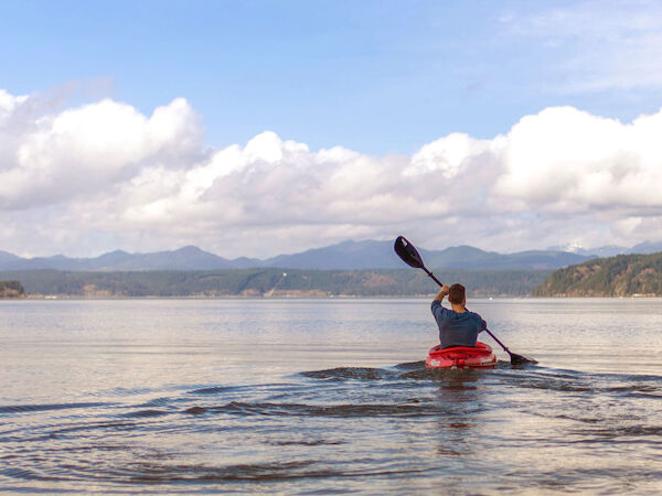 A person is kayaking on a calm lake with mountains and clouds in the background, under a bright blue sky.