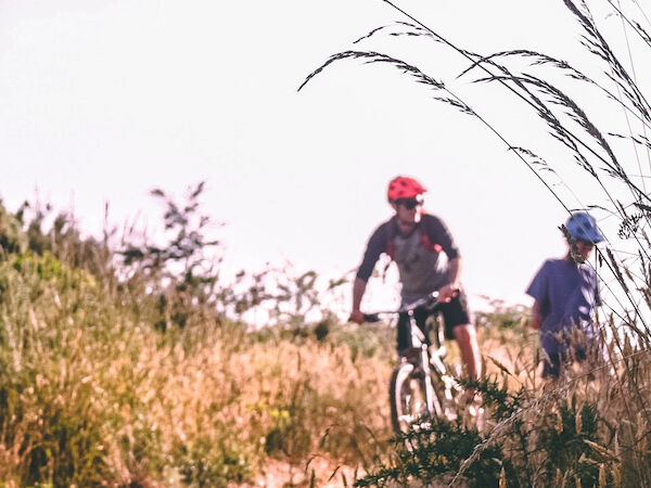 Two people are biking on a dirt path through tall grass under a clear sky, both wearing helmets.